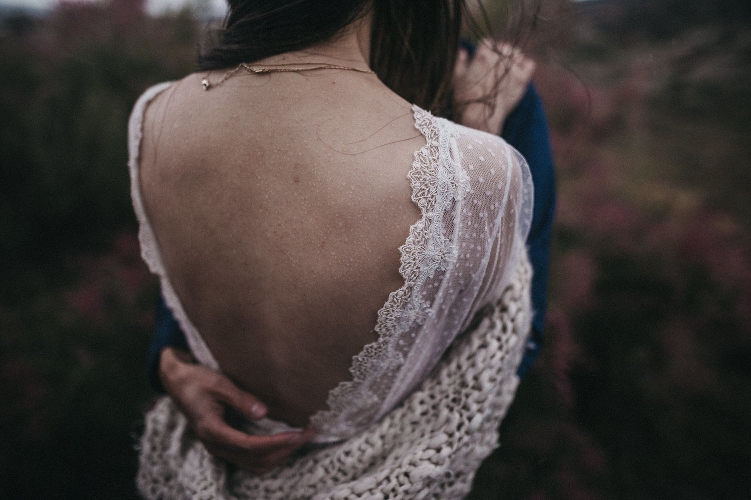 Close-up of lace wedding dress back detail with couple embracing in background.