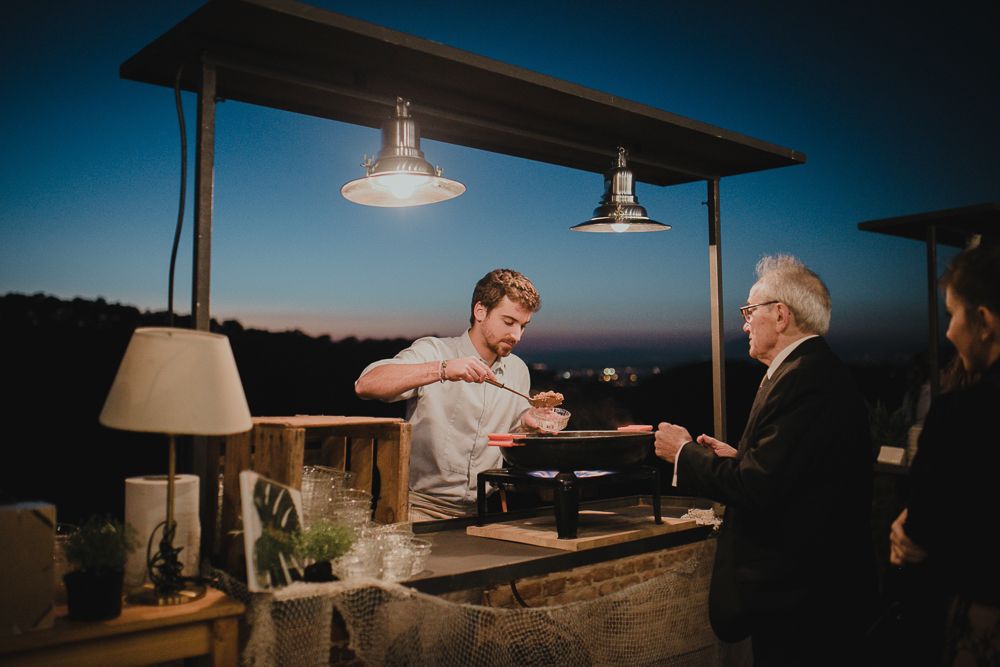 A bartender serves drinks at an outdoor bar under pendant lights at dusk with a dark blue sky in the background.
