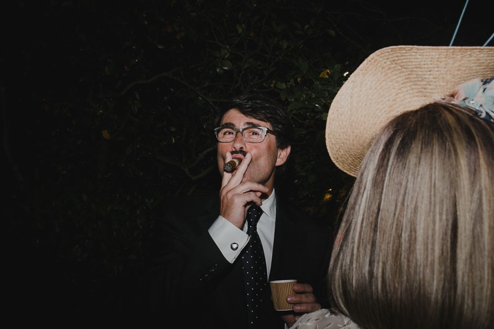 Guest in formal attire smoking a cigarette while wearing a wide-brimmed hat at a party.