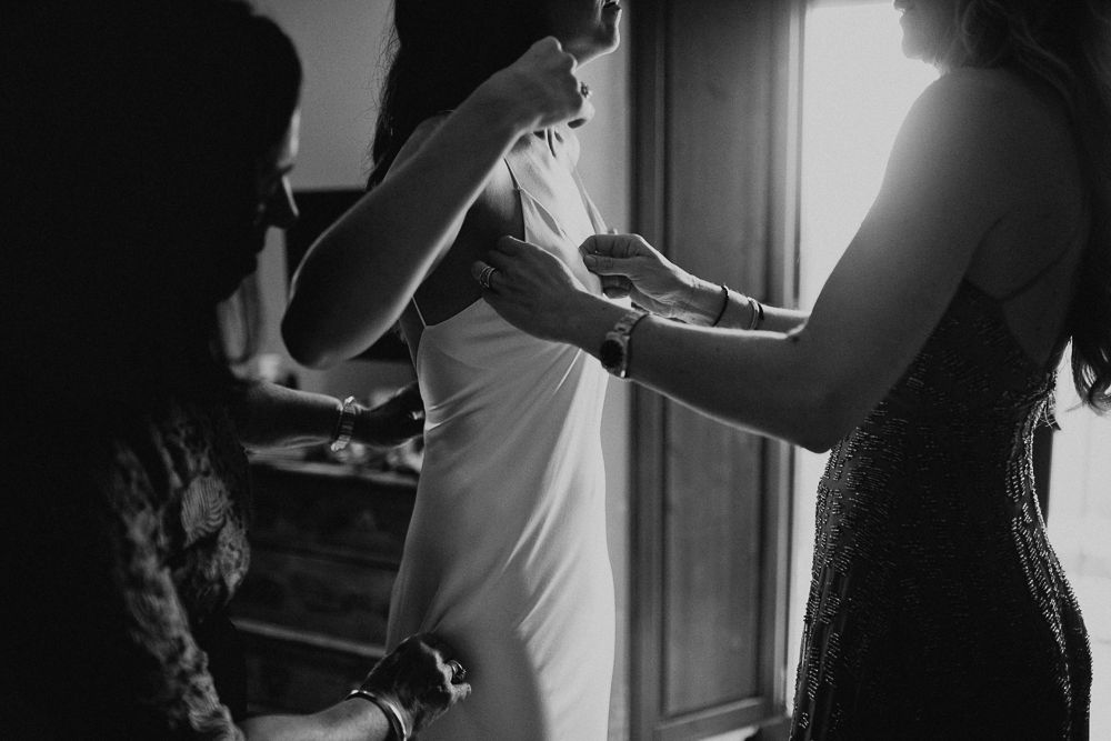 Black and white candid shot of wedding dress preparation with multiple helpers.