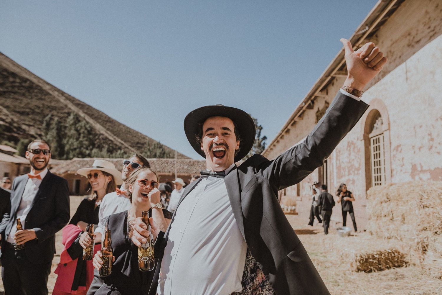 Wedding guest celebrates joyfully with arms raised outside historic stone building in desert landscape.