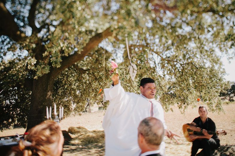 A celebrant raises their hands during an outdoor wedding ceremony under tree branches.