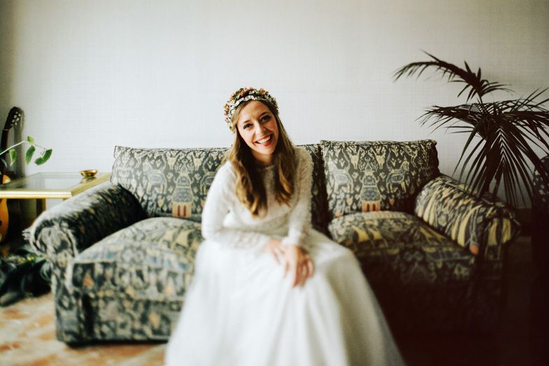 A bride in a long-sleeved white gown sits on a floral patterned couch with indoor plants.
