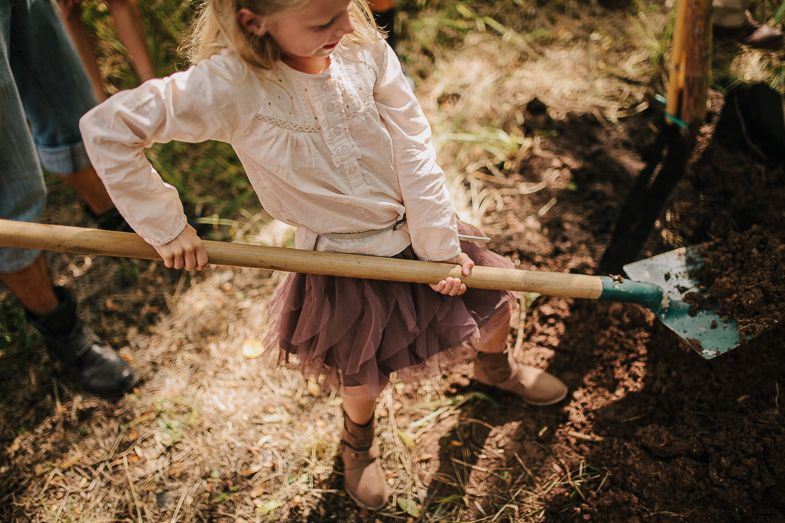 A child in a white shirt and brown skirt holds a garden tool while playing outdoors in dappled sunlight.