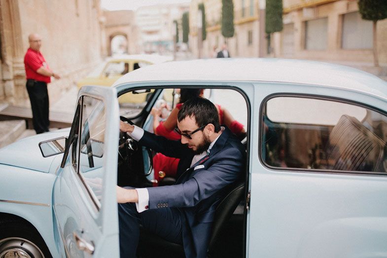 A vintage light blue car parked on a historic European street with stone architecture visible in the background.