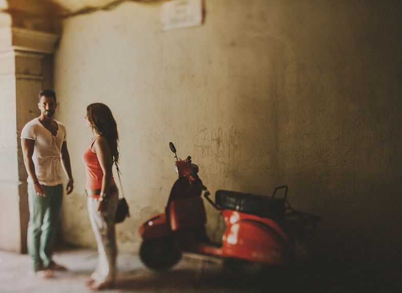 A vintage red scooter parked against a weathered wall with two people standing nearby.