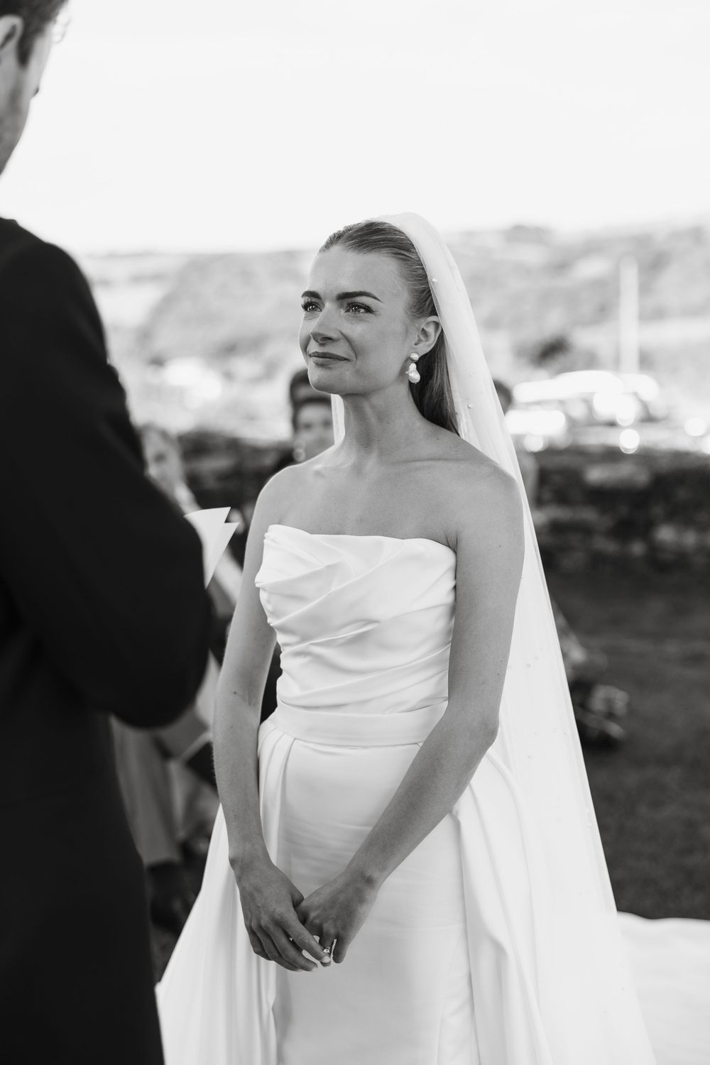 Elegant bride in classic white wedding dress with flowing veil against outdoor backdrop.