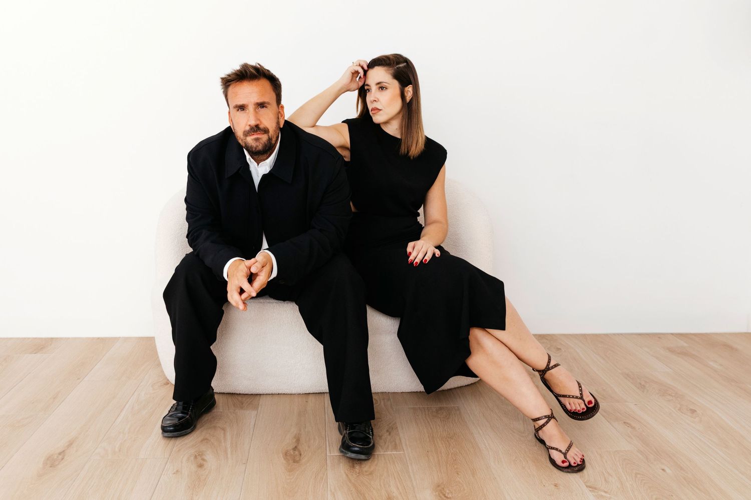 A couple in black attire sits together on a light wooden floor in a minimalist white studio setting.