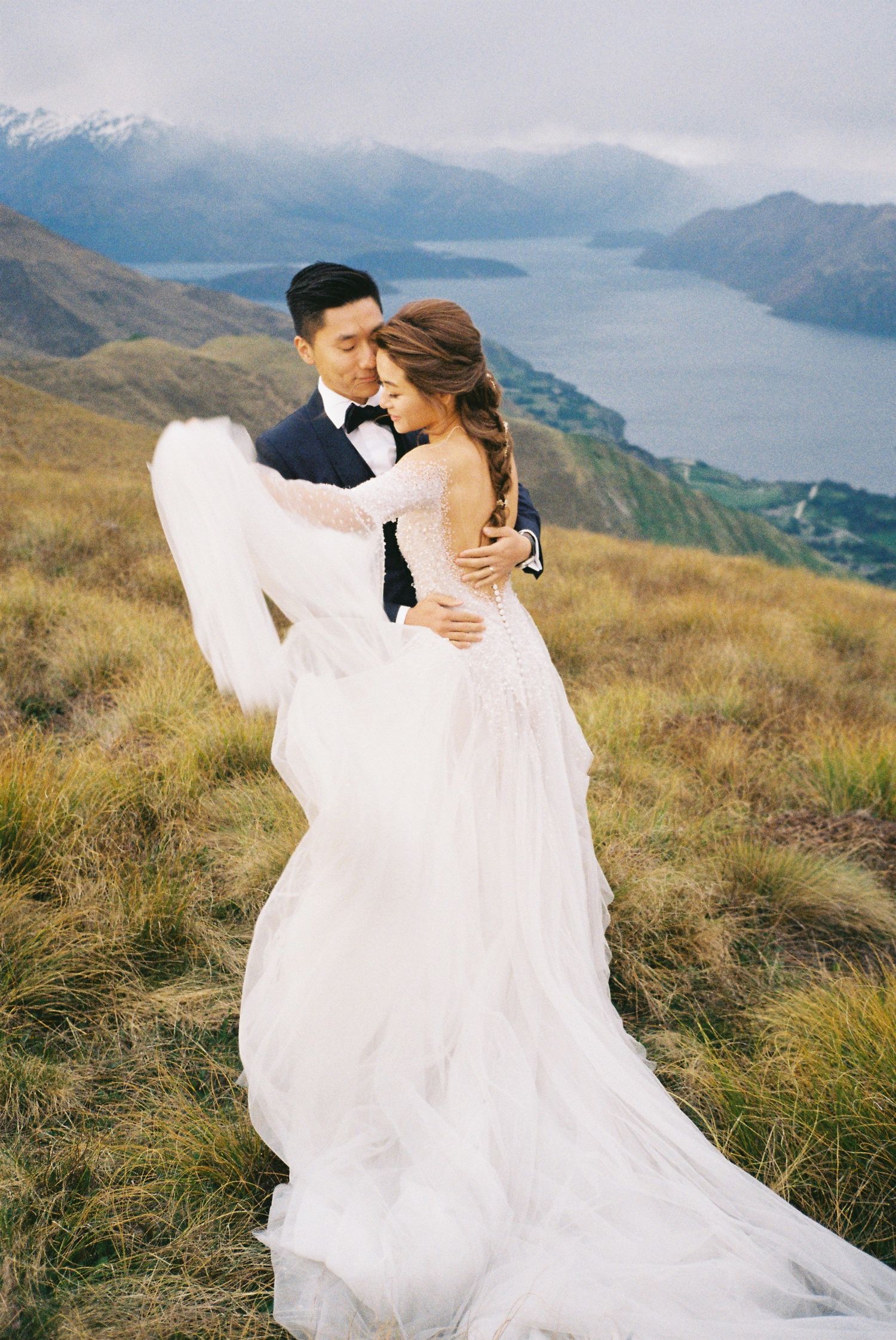 A couple embraces on a mountaintop overlooking a scenic lake and mountain landscape in New Zealand.