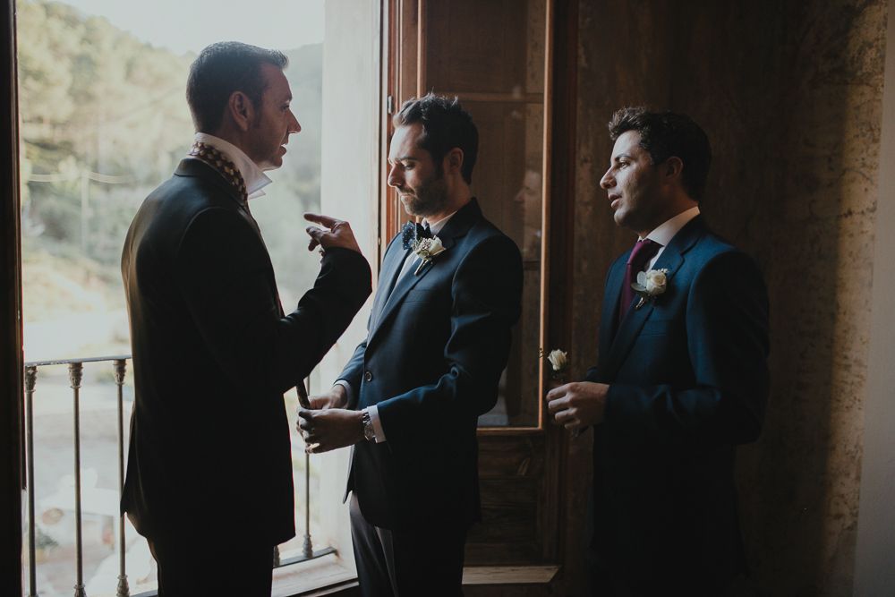 Three groomsmen in navy suits having a conversation near a window during a wedding.