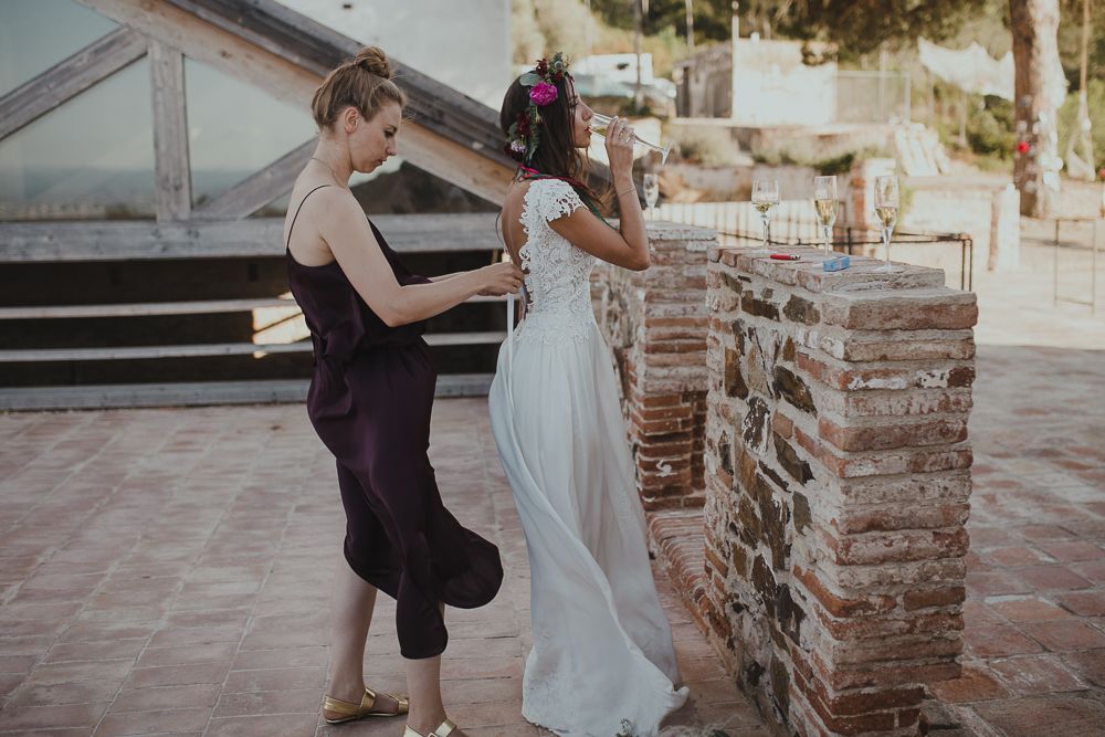 Wedding preparations take place outdoors as someone helps adjust a flowing white dress by a stone pillar in warm sunlight.