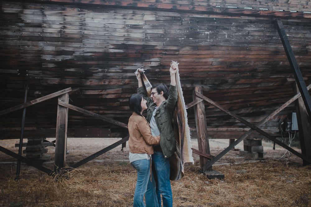 A couple shares a romantic moment against a rustic wooden structure outdoors.