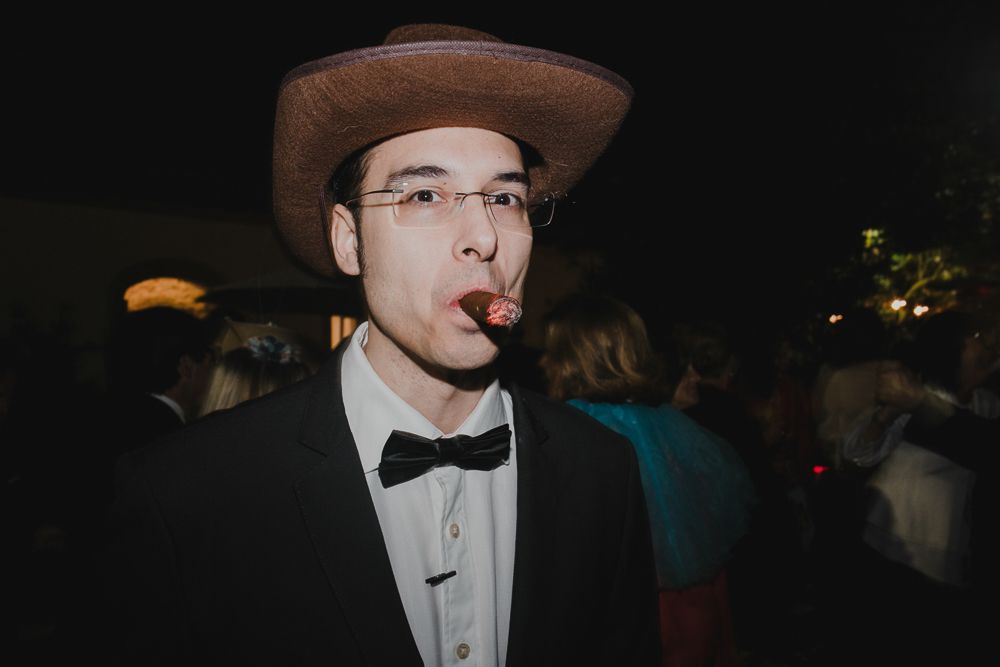 Person in formal attire with a bowtie and hat smoking at a nighttime event.