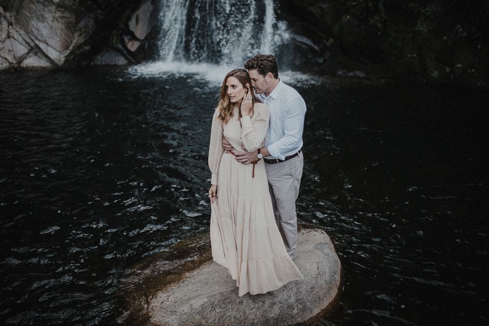A couple embraces while standing on a boulder with a misty waterfall backdrop in moody lighting.