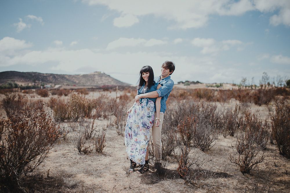 A couple poses together in a desert landscape with dry brush and blue sky.