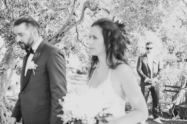 A black and white outdoor wedding photo of a couple and guest in a natural setting.
