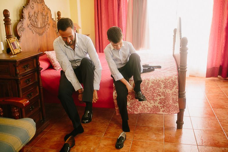 Two people putting on formal shoes while sitting on ornate red chair.