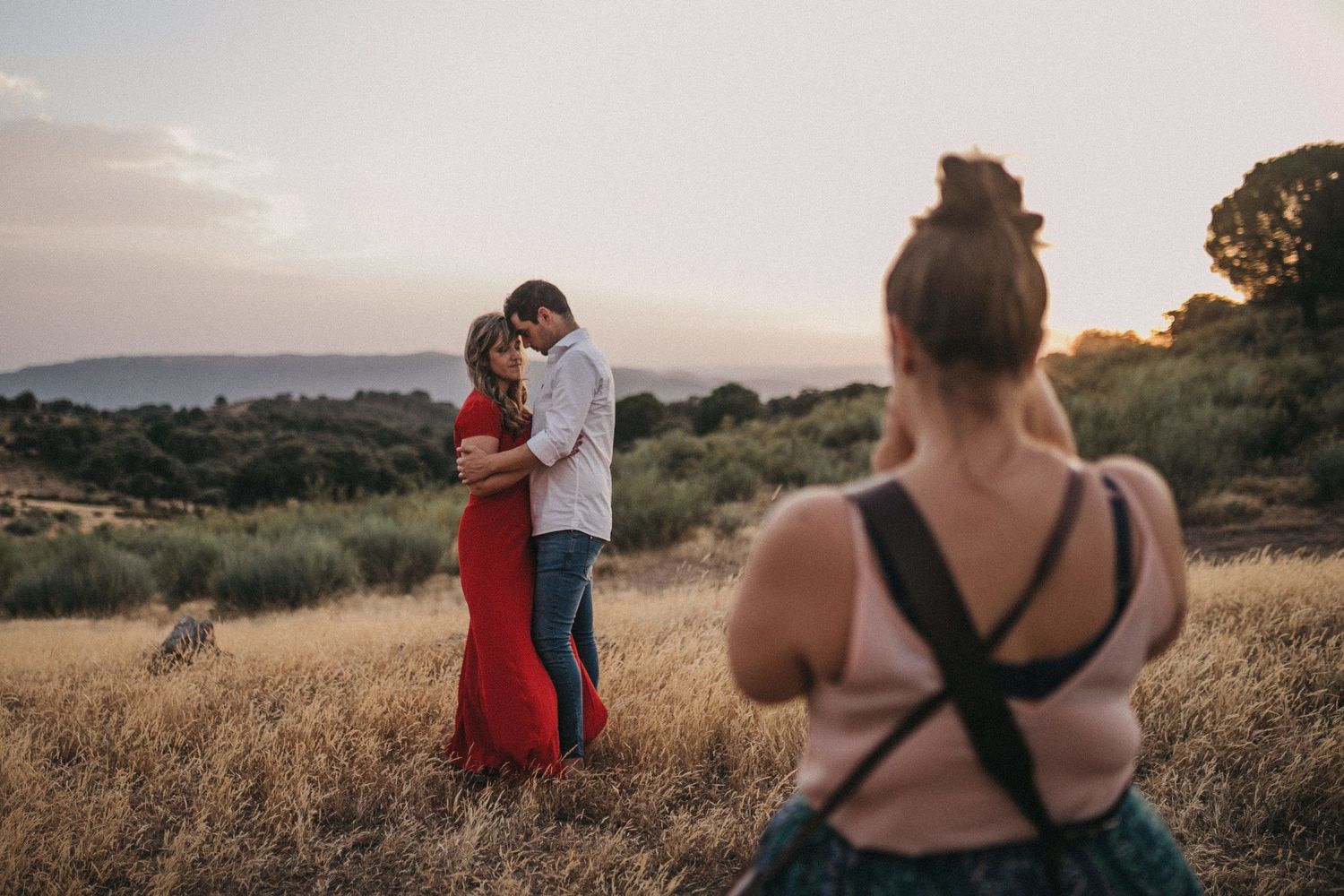 Photographer capturing couple in red dress embracing in golden field at sunset.