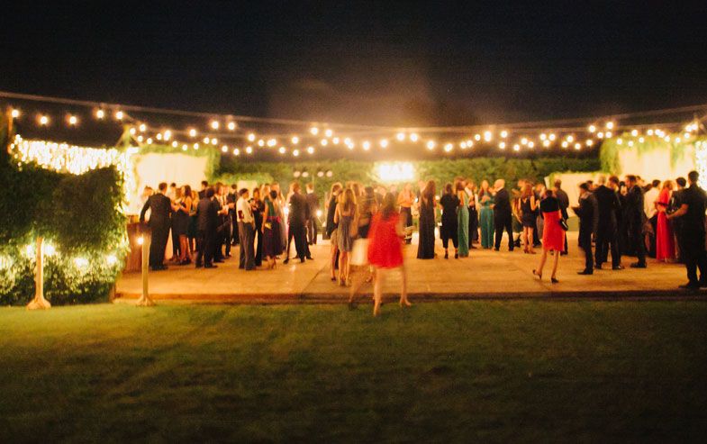 Wedding guests gather under string lights at an outdoor evening reception.