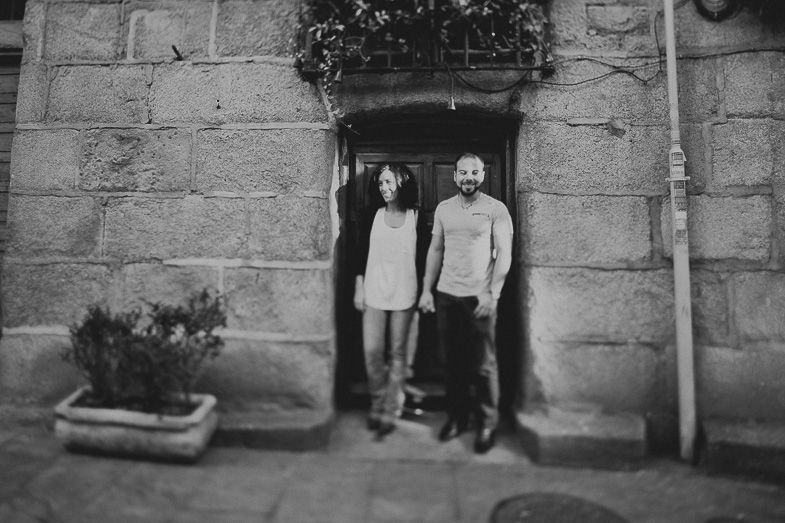 A couple stands in a rustic stone doorway with a flower planter beside them in black and white.