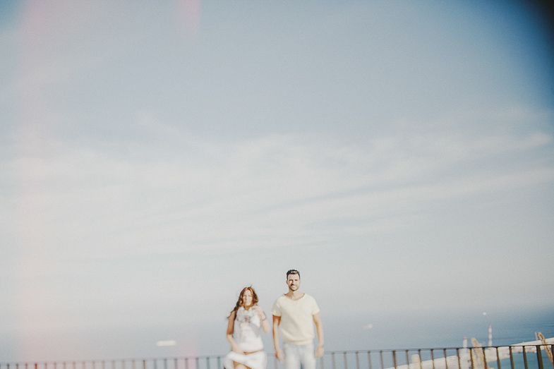 A couple in light clothing poses against a bright sky with ocean views.