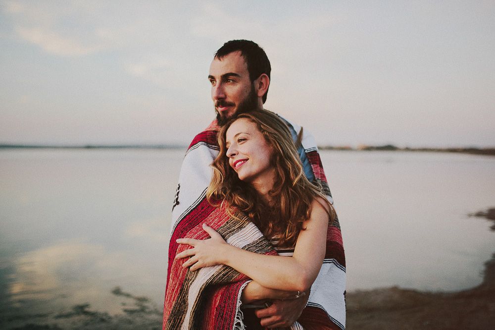 Couple embraces while wrapped in blanket at sunset by peaceful waterfront.