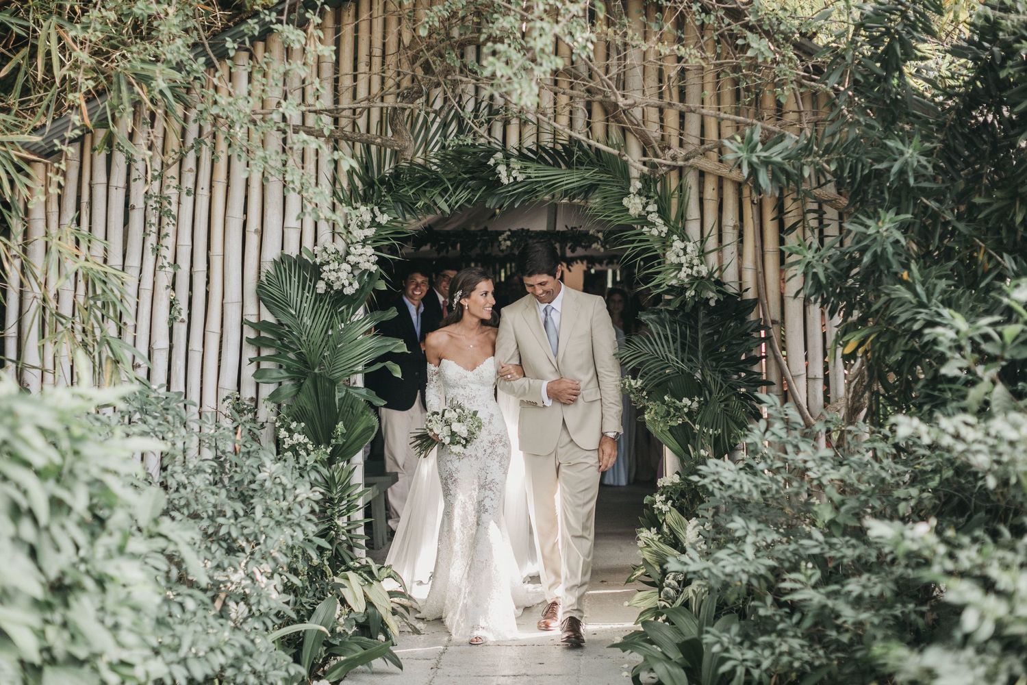 A wedding couple walks through a lush garden archway decorated with bamboo and greenery.