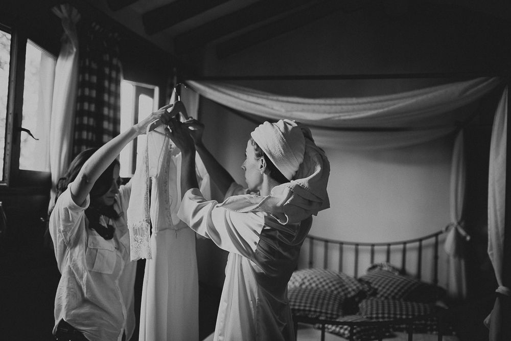 Two people help each other get ready in a bedroom with draped fabric and dark wooden furniture.