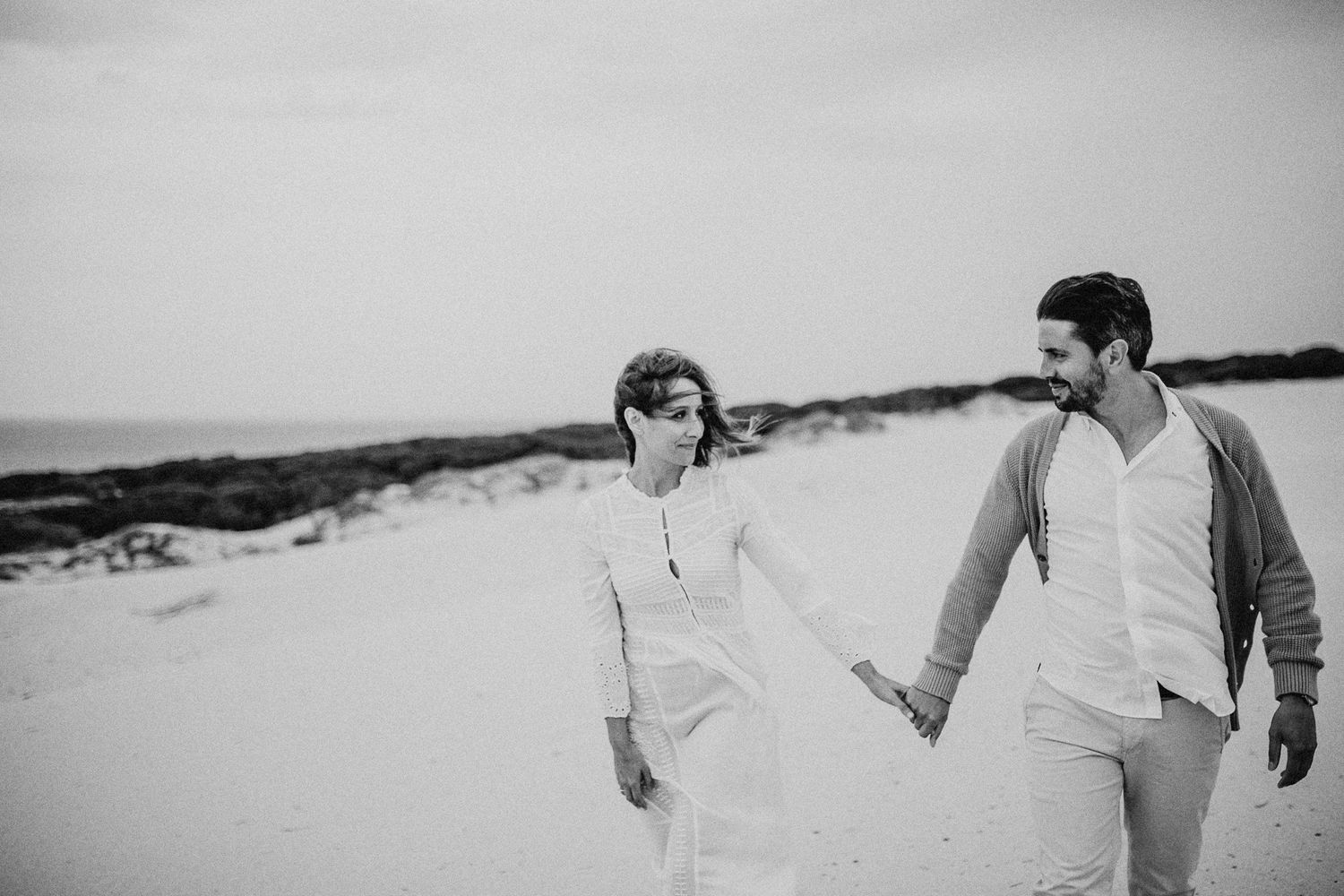 Black and white photo of couple walking hand in hand across sandy landscape.