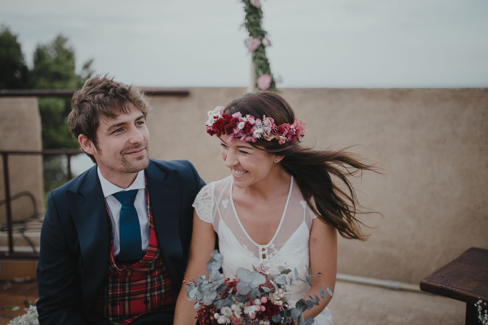 A couple in wedding attire share a candid moment outdoors with the bride wearing a floral crown.