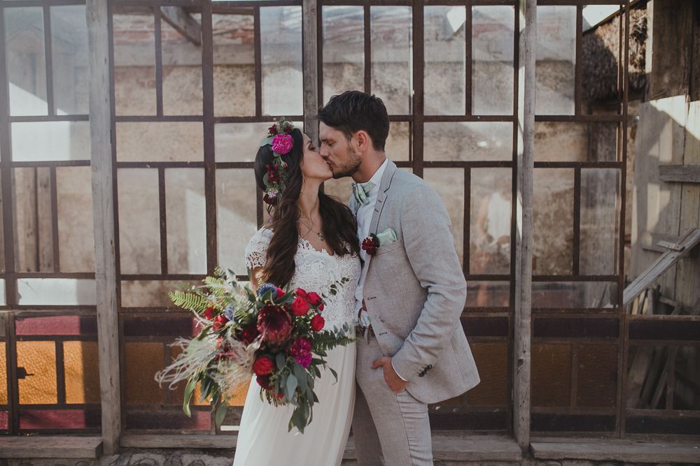 Wedding couple with vibrant bouquet posing against rustic glass window backdrop.