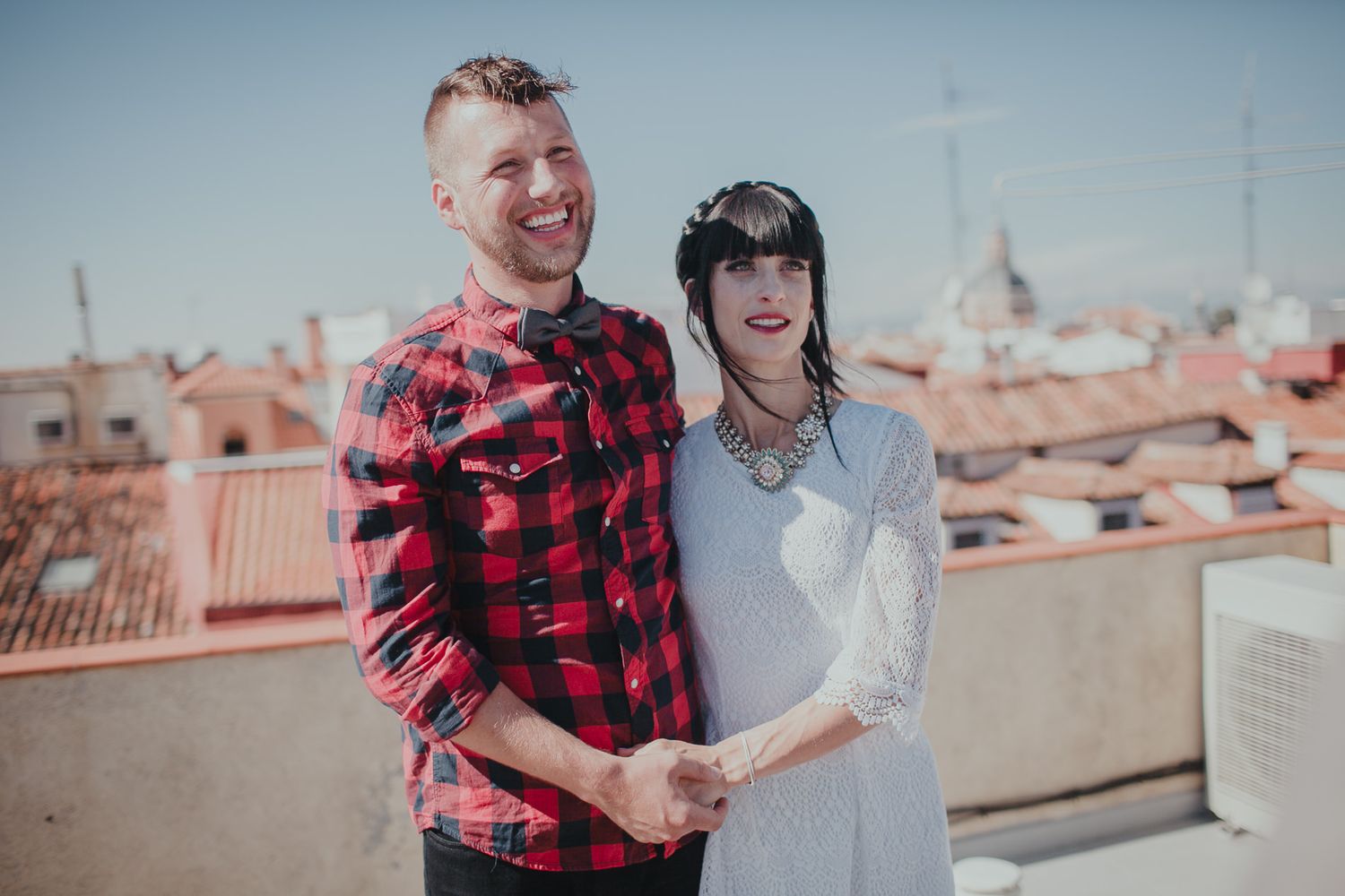 A couple posing together on a sunny rooftop with Mediterranean architecture behind them.
