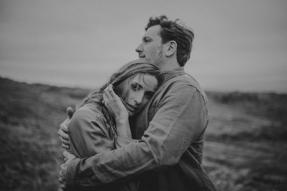 A couple embraces tenderly in a moody black and white portrait on a hillside.