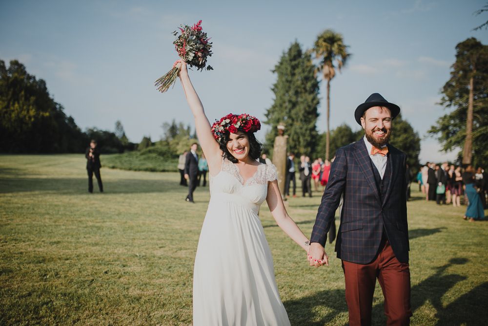 A joyful outdoor wedding celebration with red floral crown and bouquet against lush greenery.