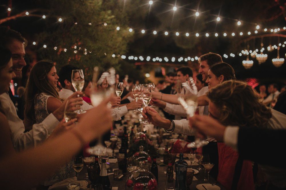 Guests raise glasses in a toast during an outdoor dinner celebration under twinkling string lights.