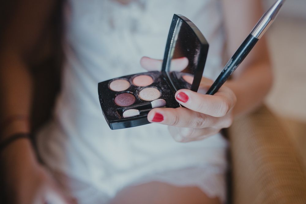 Hands holding eye shadow palette and makeup brush with red nail polish against blurred background.