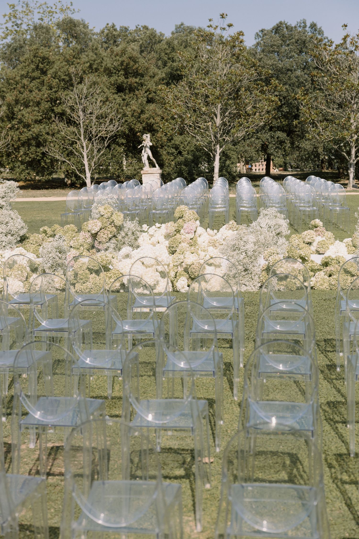 Clear acrylic chairs and white floral arrangements set up outdoors with trees and a statue in the background.