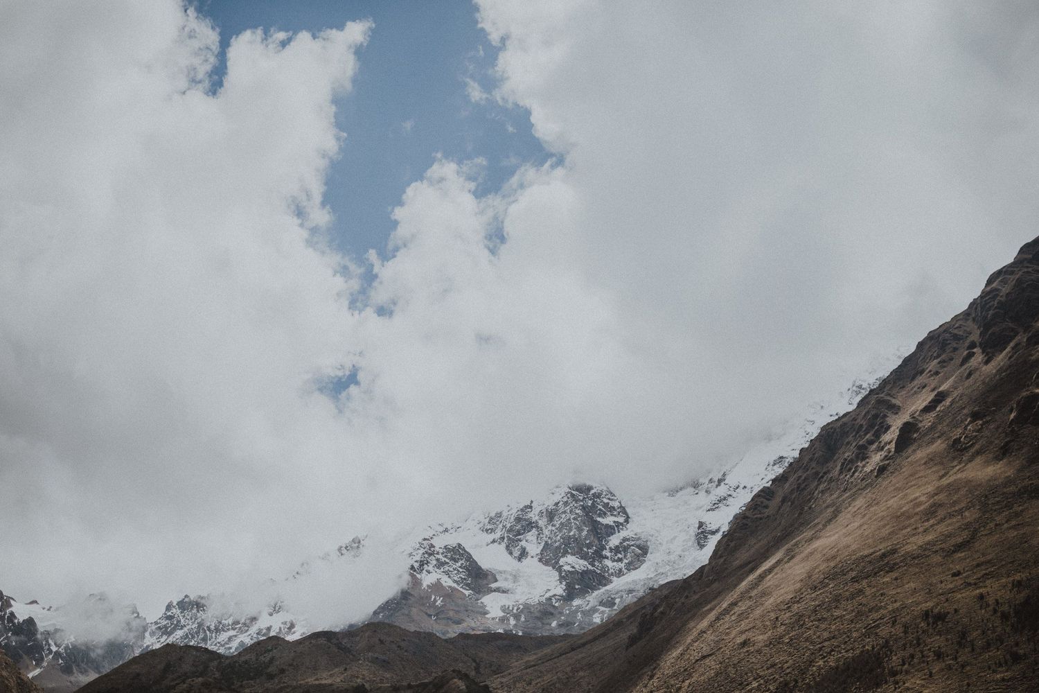 Snow-capped mountain peaks partially obscured by dramatic clouds in rugged terrain.