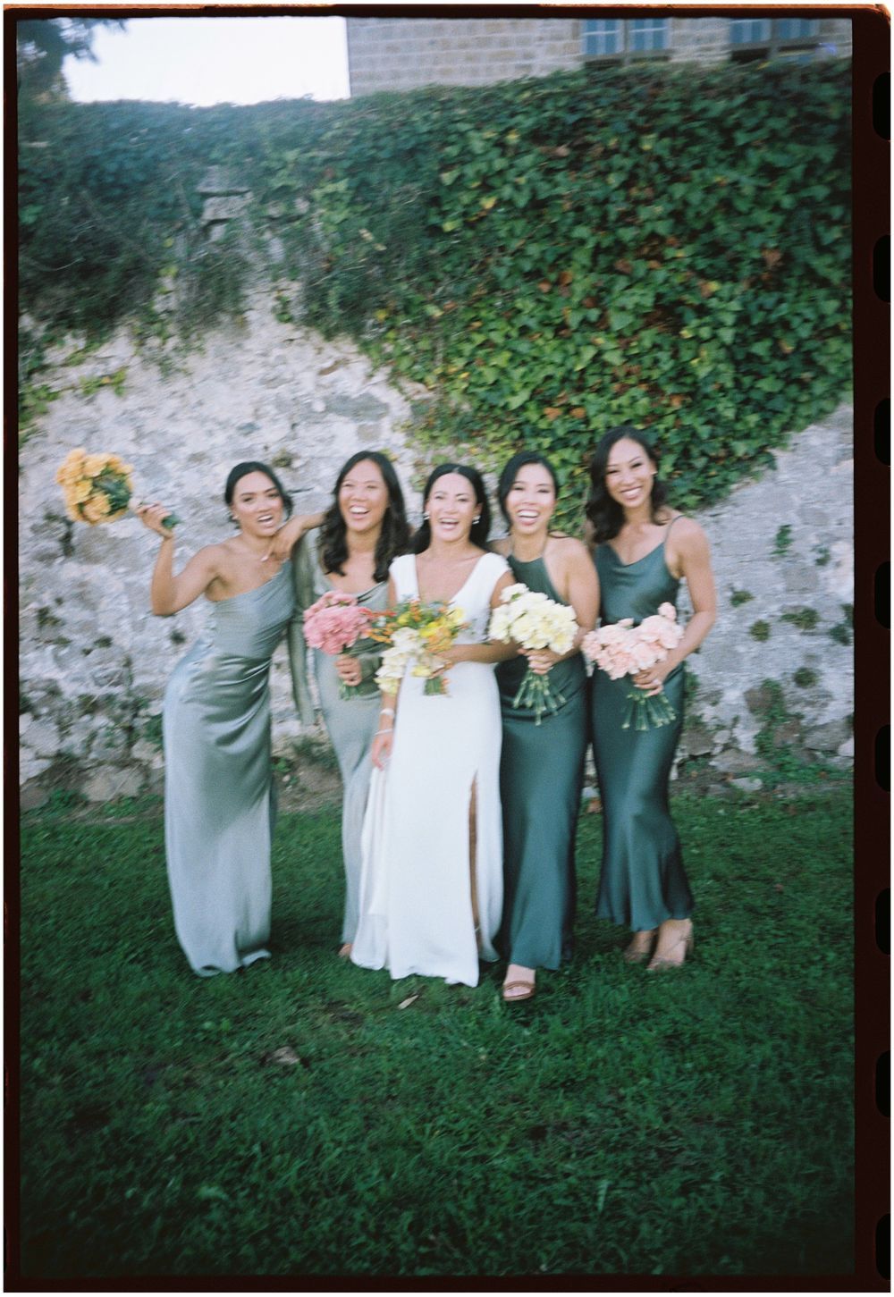 Wedding party in coordinated grey dresses posing against an ivy-covered wall.