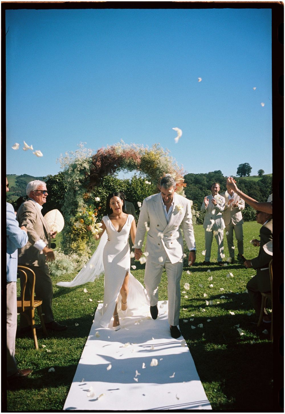 Outdoor wedding celebration with white flower arch and guests throwing petals.