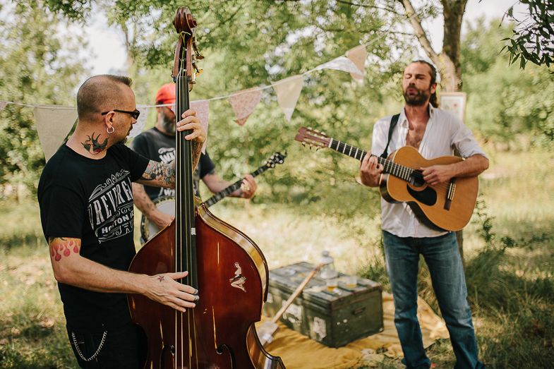 Two musicians perform outdoors with an upright bass and acoustic guitar in a sunny woodland setting.