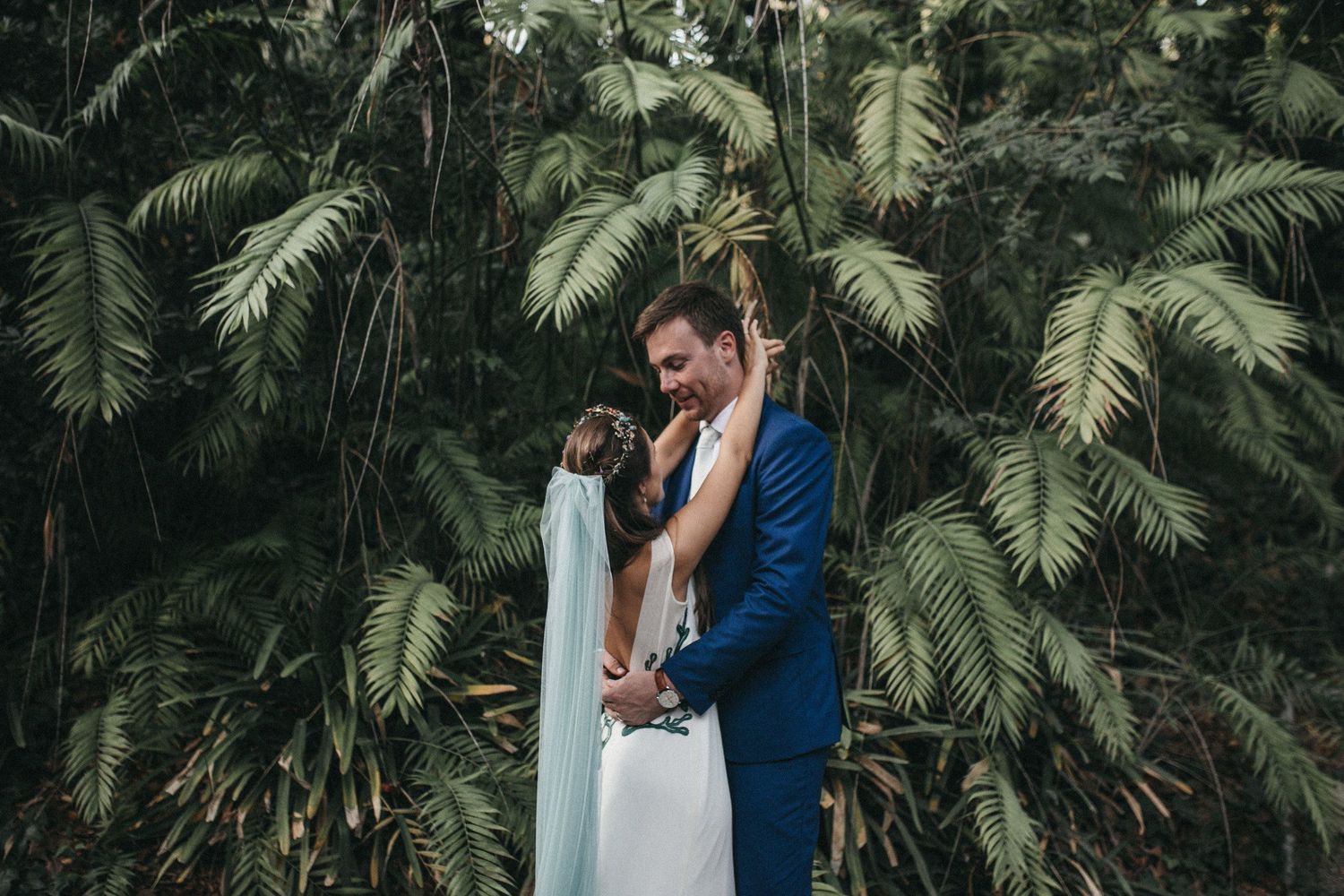 A couple embraces amidst lush tropical palm fronds during their wedding photoshoot.