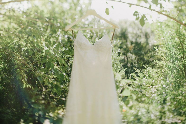 A delicate white wedding dress hangs outdoors among green leaves and natural lighting.