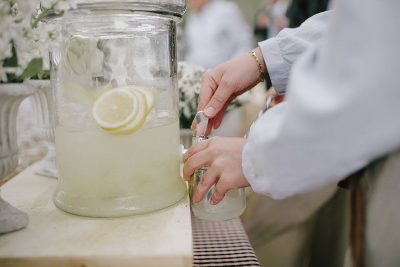 Hands pouring refreshing lemonade from a glass beverage dispenser.