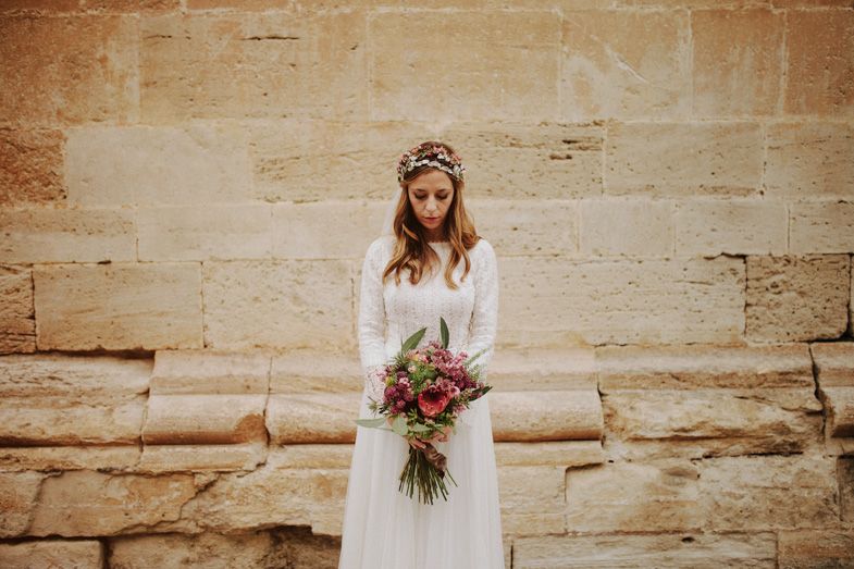 Bride in vintage-style dress holds colorful bouquet against stone wall backdrop.