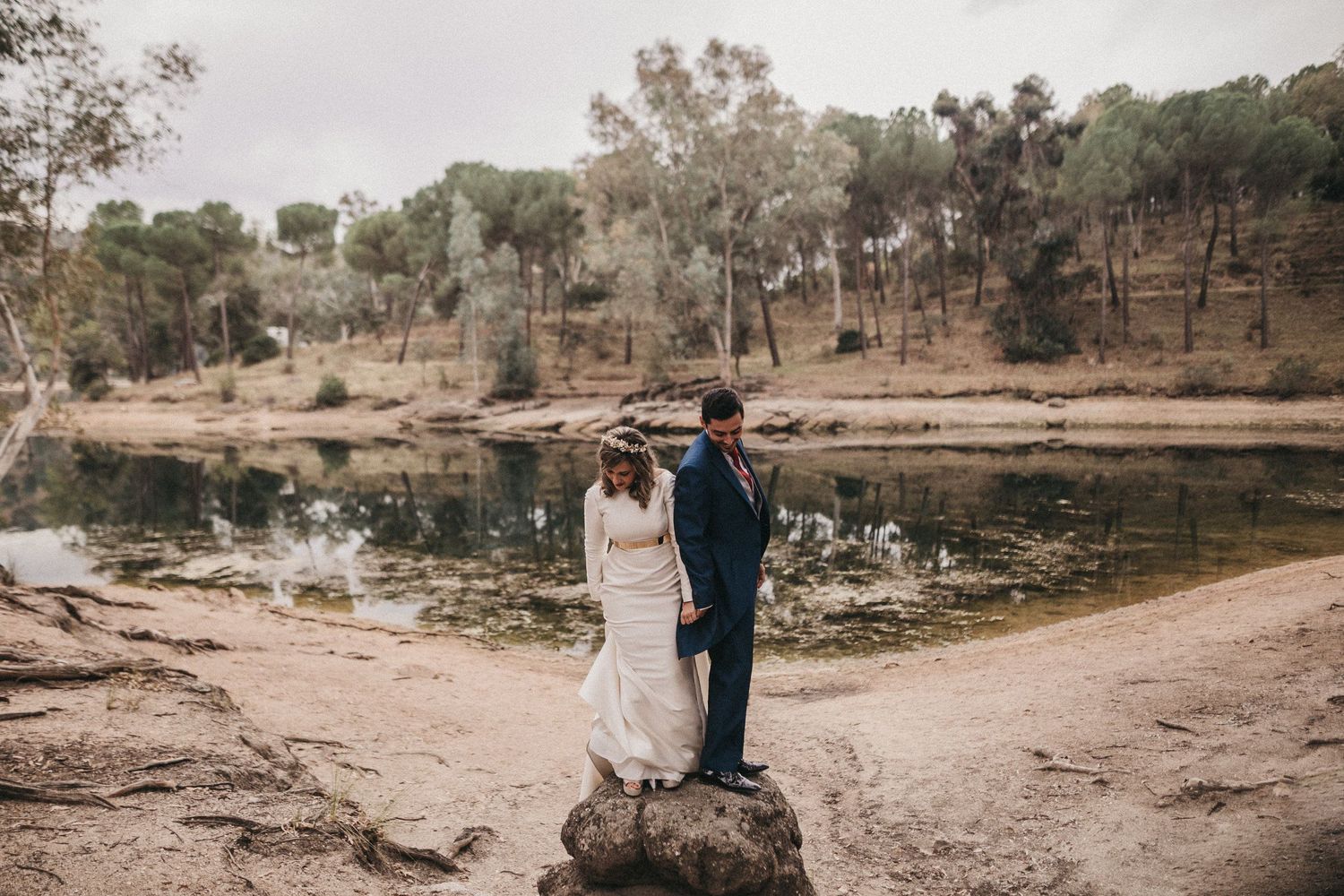 A wedding couple stands together on rocks overlooking a serene lake surrounded by trees.