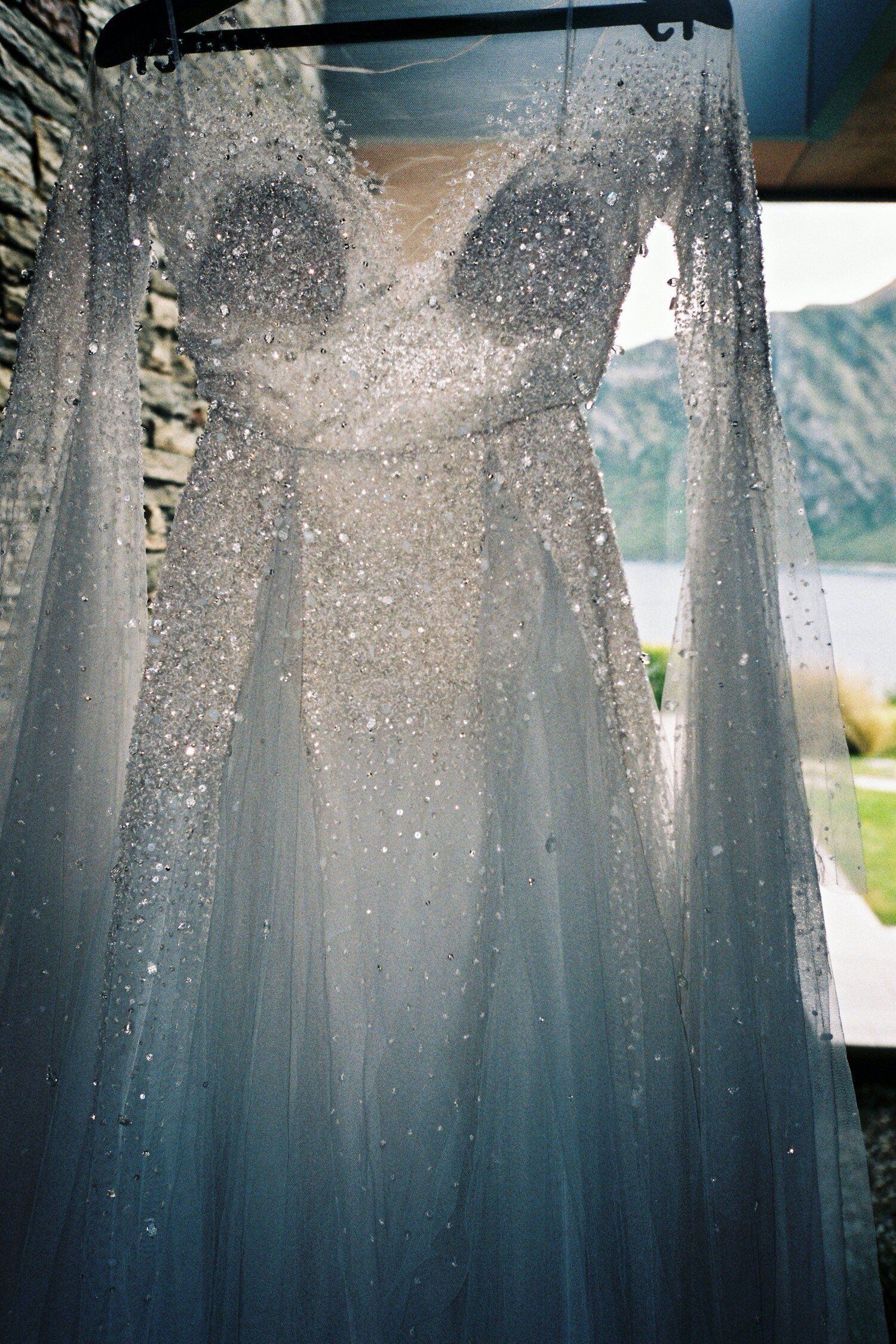 A sparkly wedding gown with intricate beading hangs against mountain landscape backdrop.