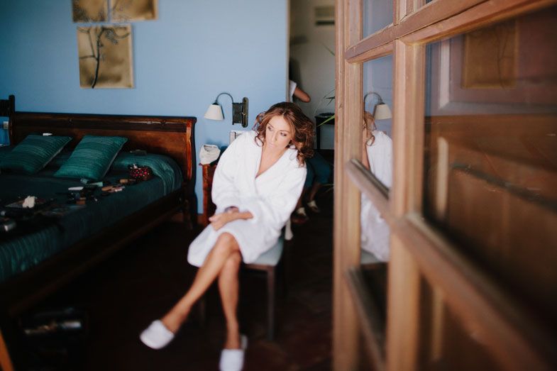 Person sitting in a white robe and sneakers in a dimly lit room with wooden furniture.
