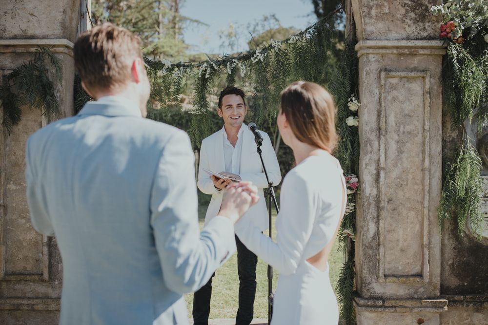 An officiant conducts a wedding ceremony beneath a stone archway decorated with floral arrangements.