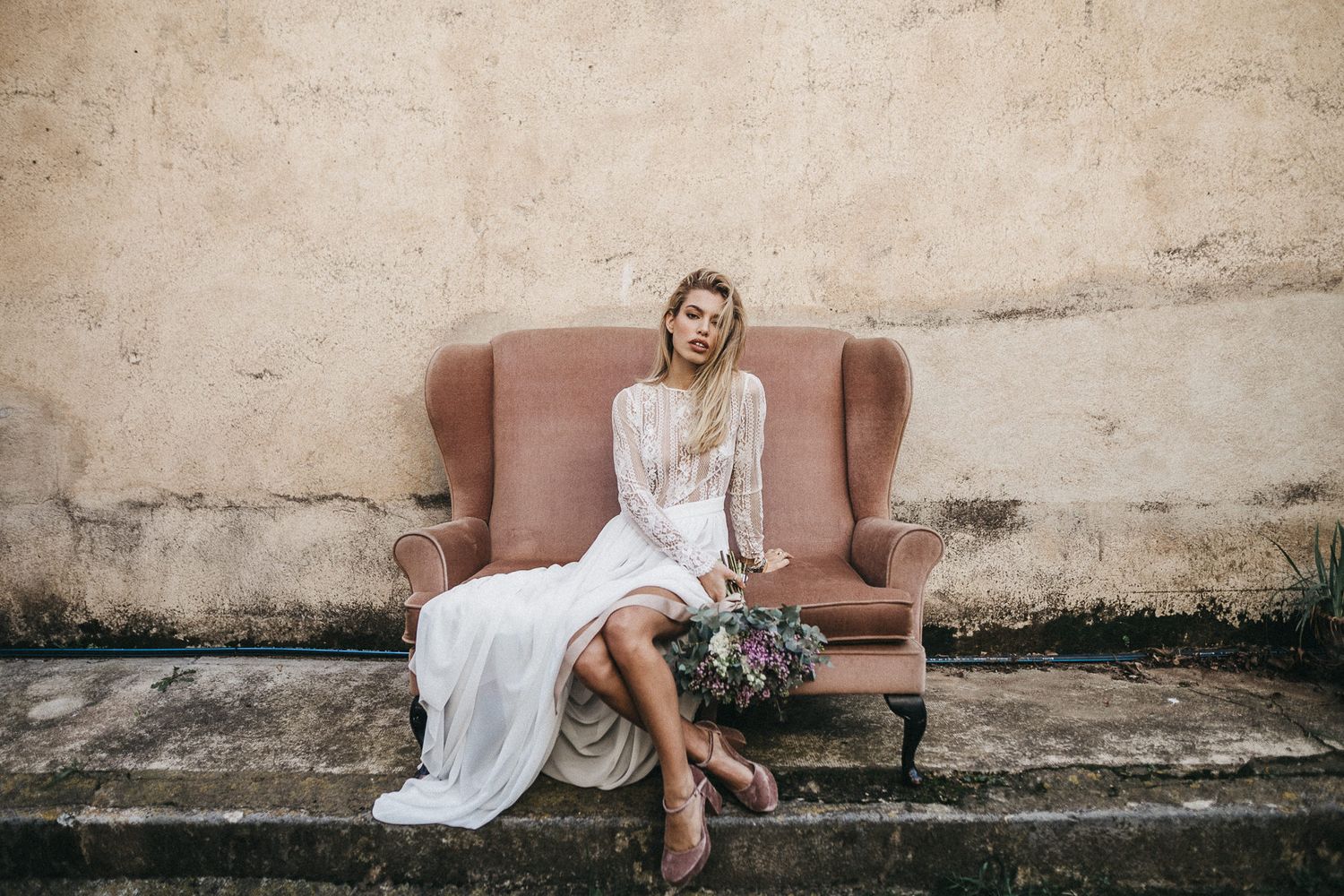 A bride in a flowing white dress sits elegantly on a vintage sofa against a weathered wall.